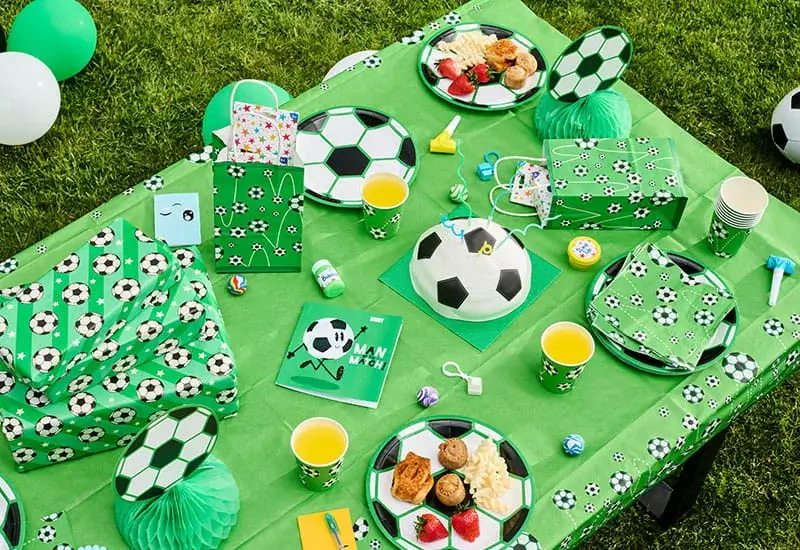 Football-themed party table with football-print tableware, a football-shaped cake, party bags, snacks, drinks, and green decorations on the grass.